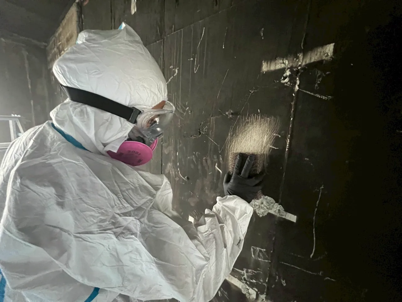 防護服の作業員が煤で黒い壁を丁寧に清掃する様子